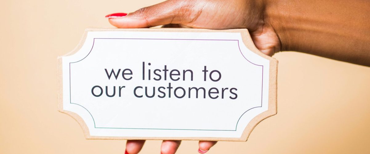 A hand holding a sign saying 'we listen to our customers' on a neutral background.